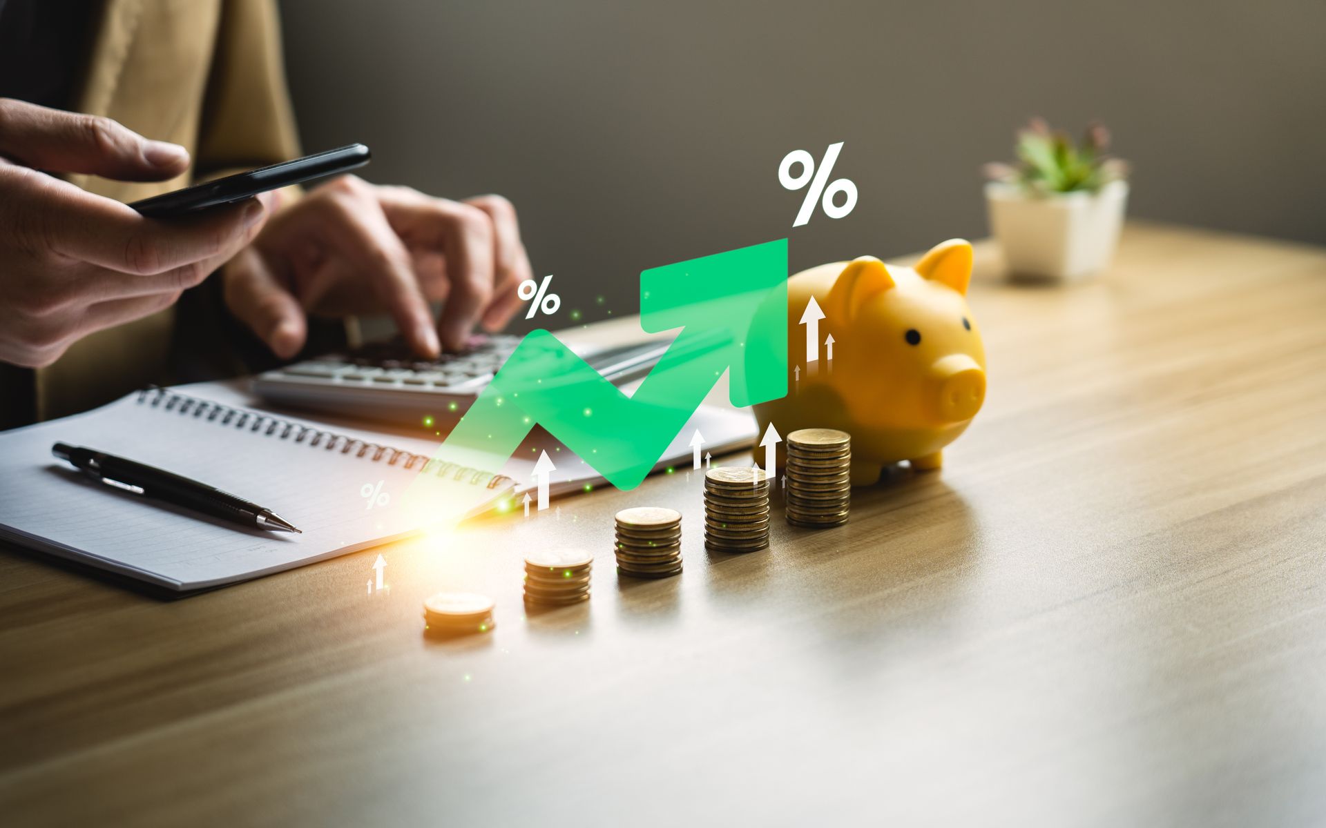 Hands using smartphone beside laptop, rising green graph, stacked coins, and a yellow piggy bank on desk