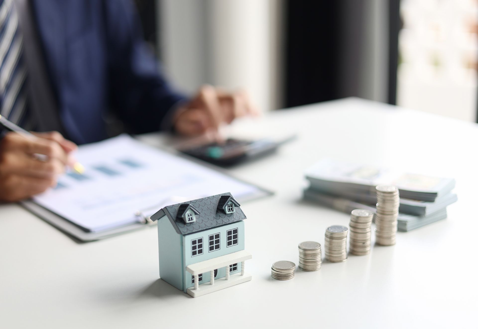 Miniature house beside stacked coins on a desk, with a person reviewing paperwork in the background