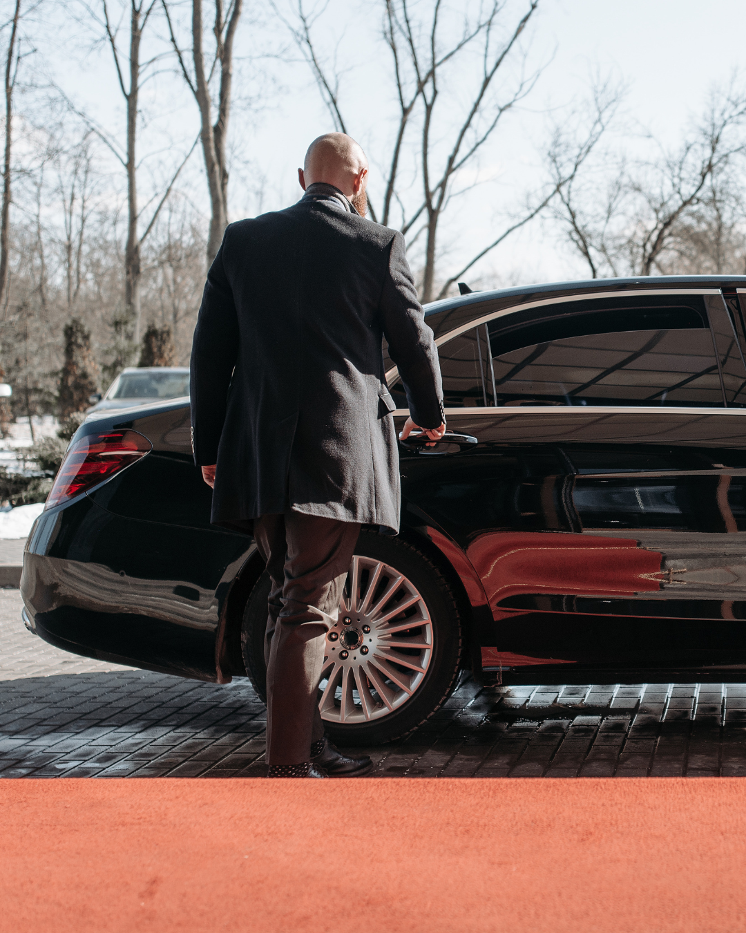 Un homme en costume sombre monte dans une berline noire, sur un tapis rouge, à l'extérieur.