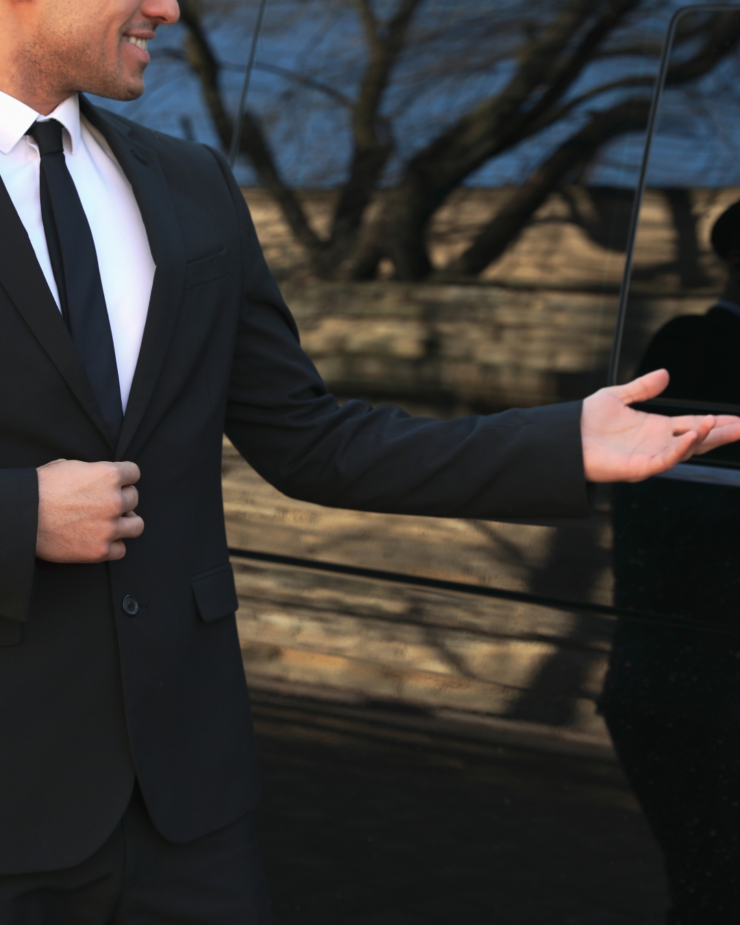 Un homme en costume désigne une voiture noire en souriant.