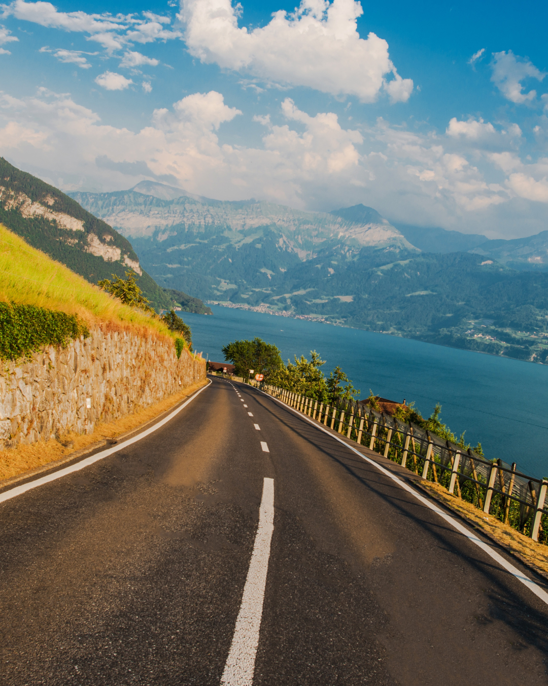 La route serpente le long d'un lac, bordée par une colline et une clôture. Au loin, on aperçoit des montagnes et des nuages.