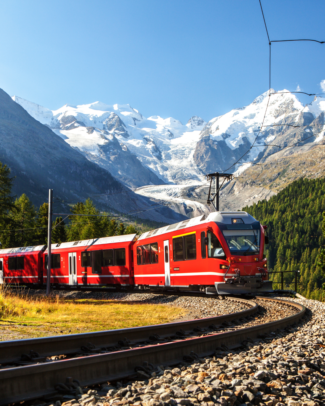 Un train rouge sur les rails décrit une courbe vers des montagnes enneigées sous un ciel bleu.