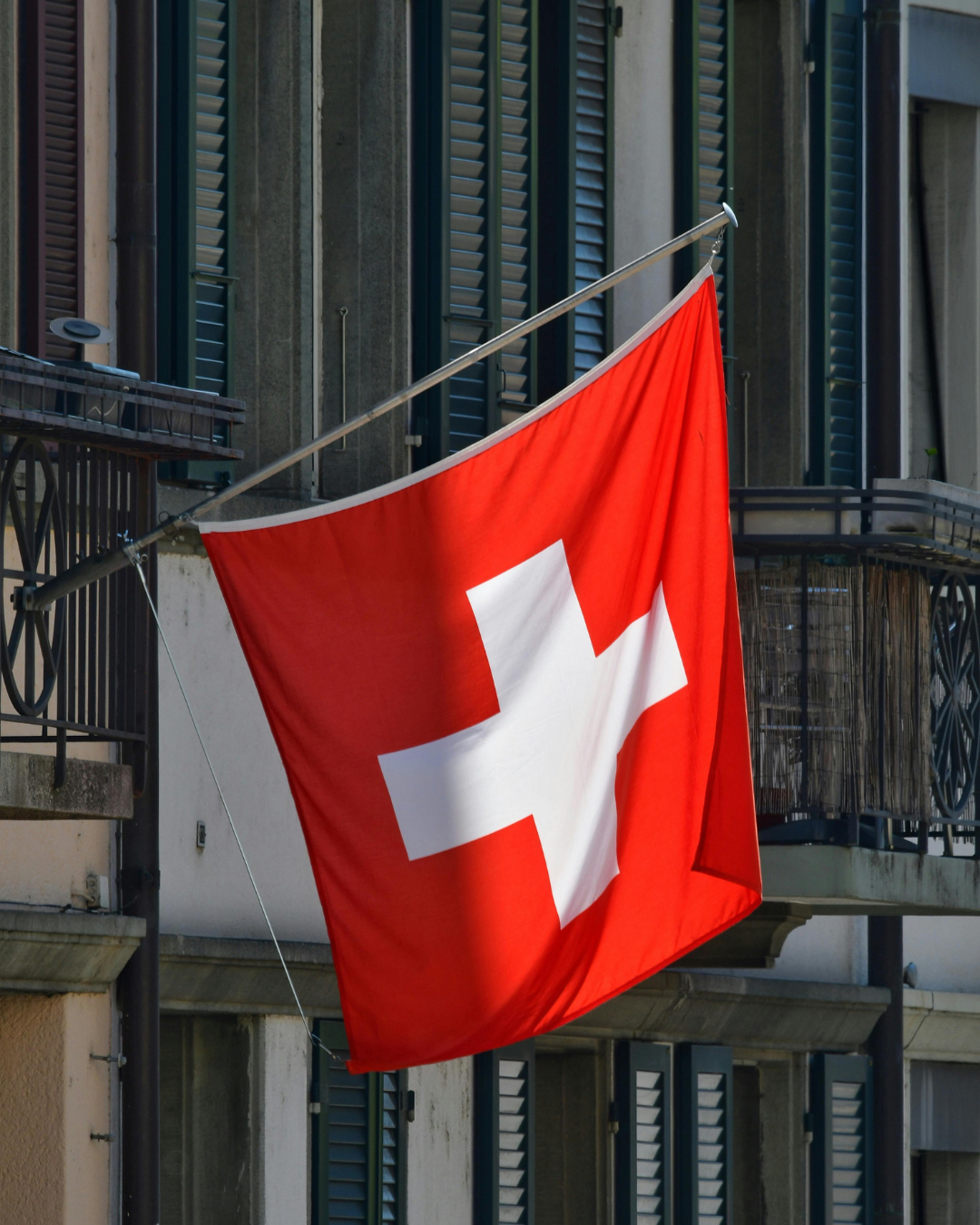 Drapeau suisse flottant devant un bâtiment à volets, fond rouge avec croix blanche.