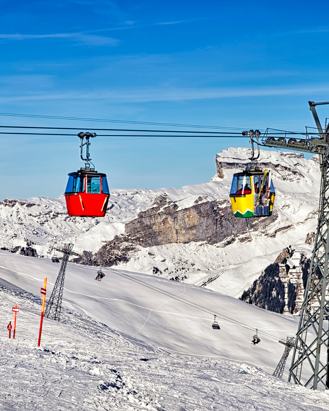 Téléphériques gravissant une montagne enneigée, ciel bleu. Cabines rouges et jaunes, skieurs visibles.
