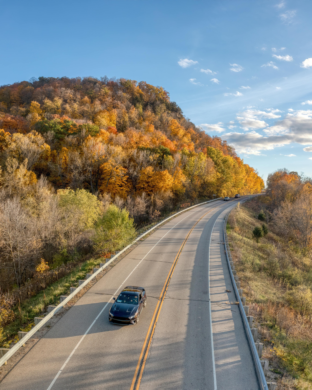 Une voiture noire roule sur une route sinueuse bordée d'arbres aux couleurs d'automne, sous un ciel bleu.