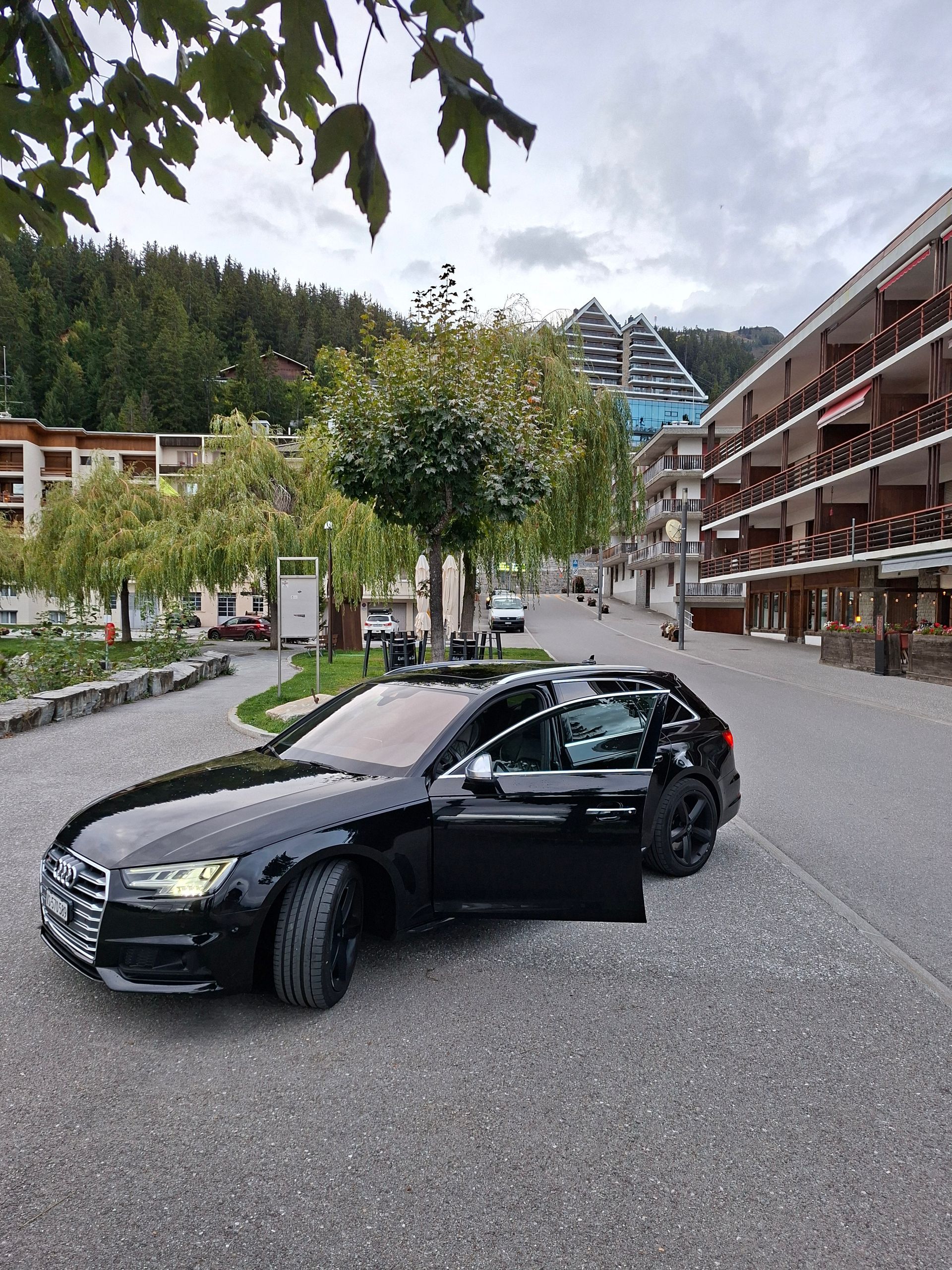 Break Audi noir garé dans une rue pavée d'une ville européenne ; bâtiments et arbres en arrière-plan.