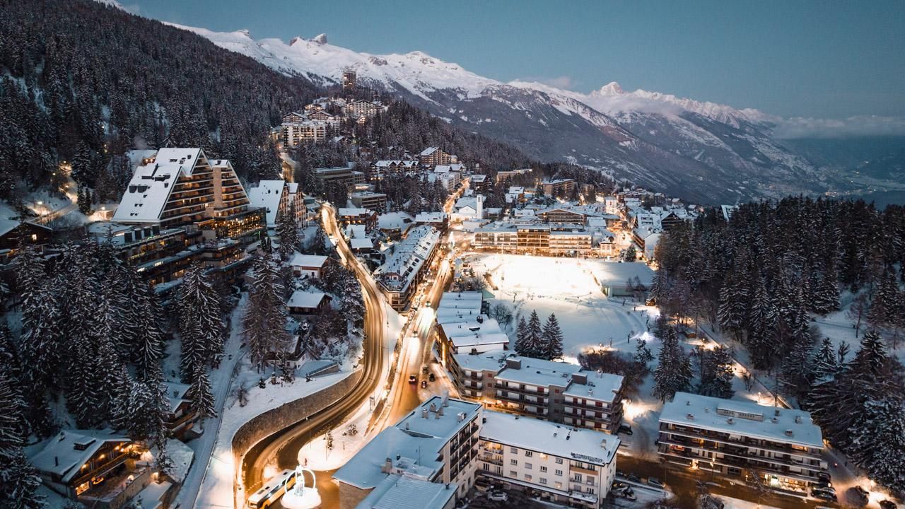 Village de montagne enneigé au crépuscule, bâtiments illuminés et routes sinueuses à travers une vallée.