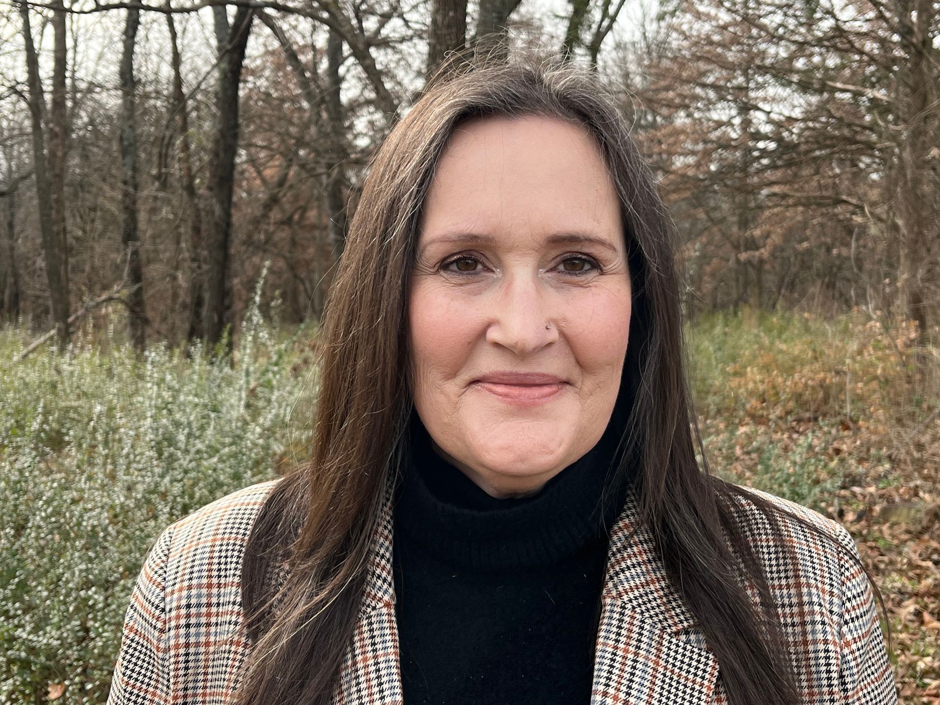 Woman with long brown hair, wearing a plaid blazer and black turtleneck, smiles in a wooded area. Anna-Lisa Johanson