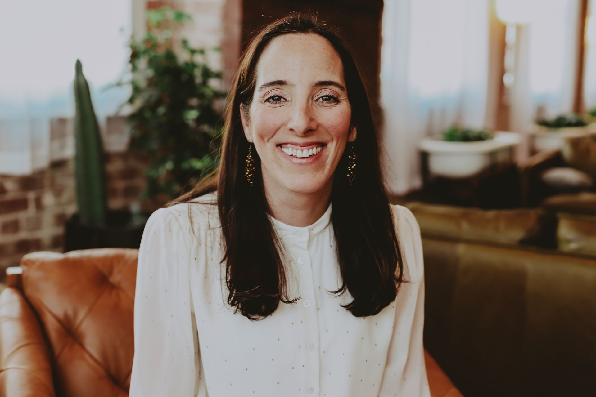 Woman with long dark hair smiles at the camera, wearing a white blouse, sitting on a leather chair. Carol Vella