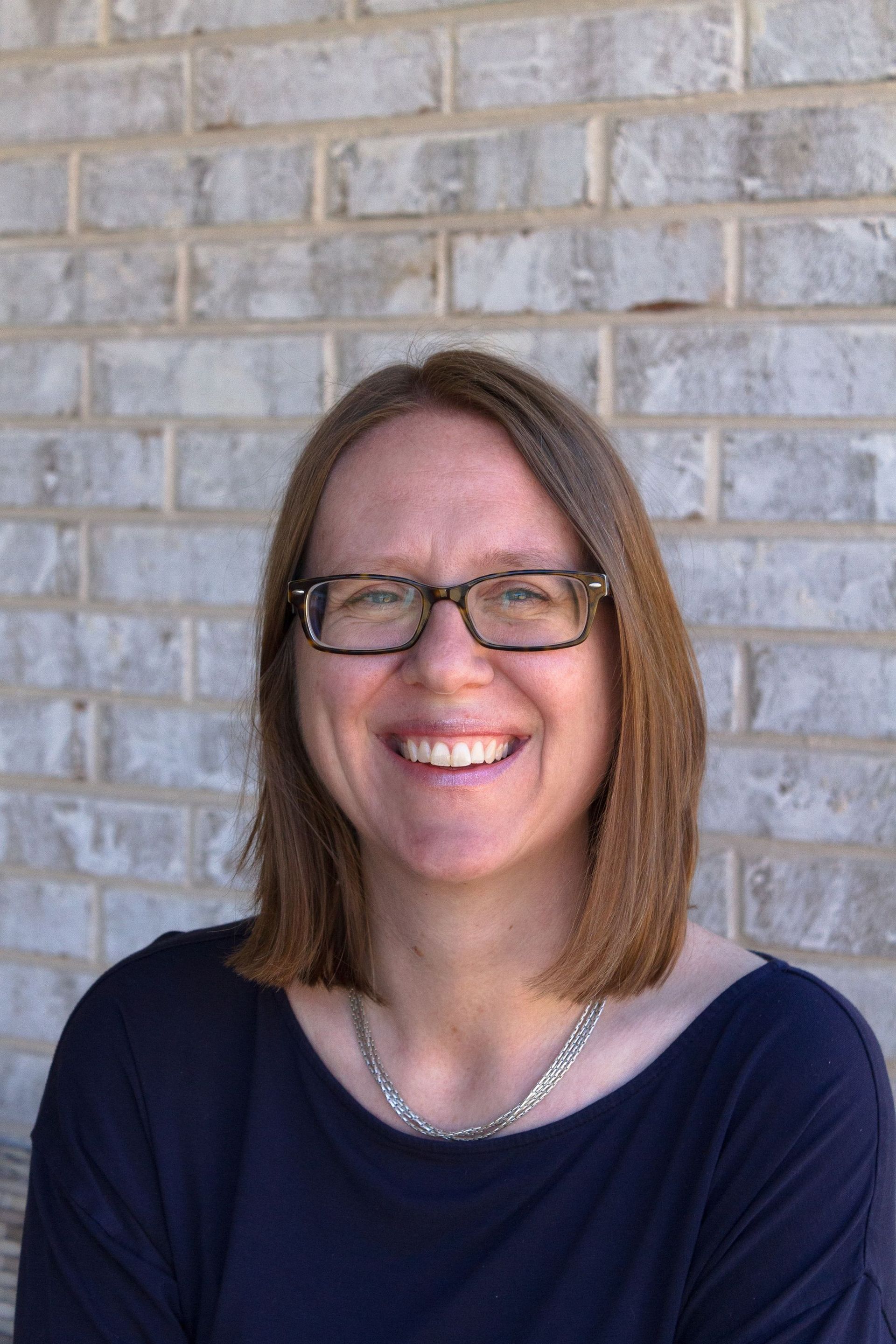 Woman with glasses smiles in front of a brick wall, wearing a dark blue top and a silver necklace. Christine Erwin-Jimenez