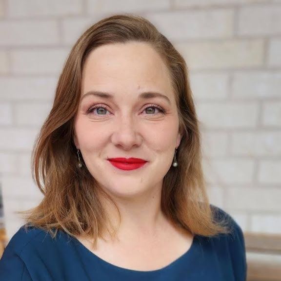 Woman with red lipstick smiles at the camera, wearing a blue shirt. Brick wall background. Katherine Tipton