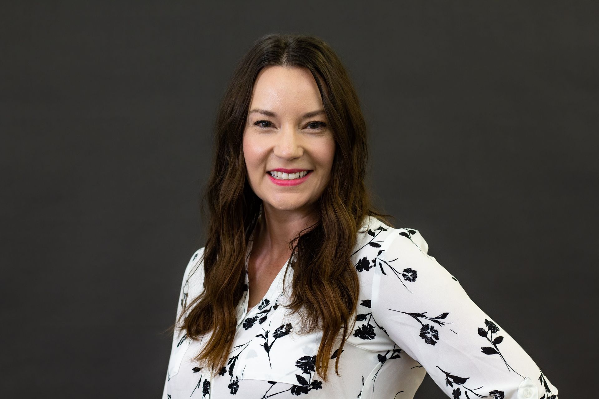 Woman with long brown hair, wearing a white floral print blouse, smiling against a gray background. Sarah Luhtanen