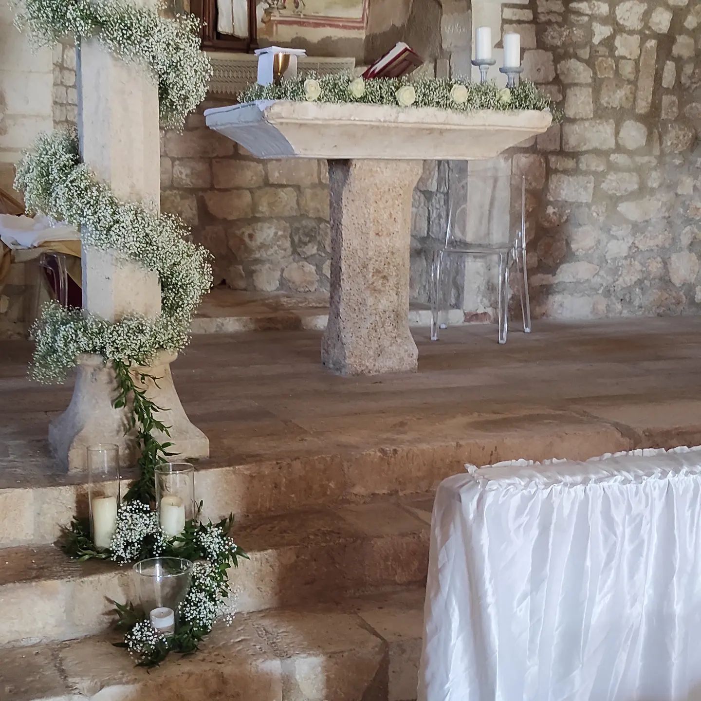 Altare in pietra decorato con fiori bianchi e verde in una cappella rustica, con un tavolo drappeggiato di bianco di fronte.