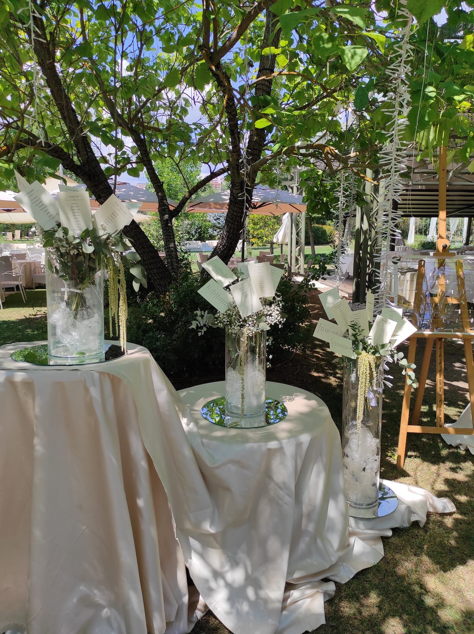 Tavolo da evento all'aperto con tovaglie bianche, vasi di vetro con fiori bianchi e una chioma di alberi soprastante.