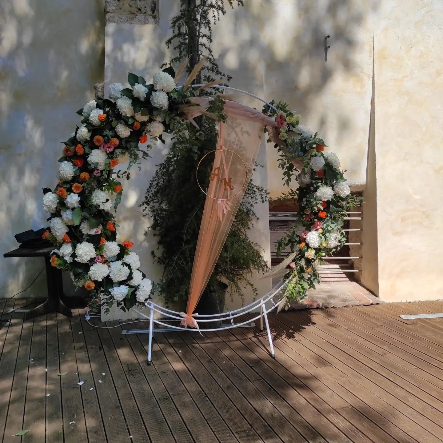 Arco floreale con fiori bianchi, arancioni e verdi su una terrazza in legno accanto a scale e un muro
