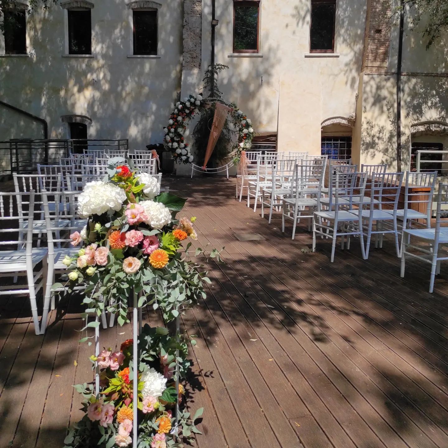 Allestimento per cerimonia nuziale all'aperto con sedie bianche, decorazioni floreali lungo la navata e un arco di sfondo su una terrazza in legno.