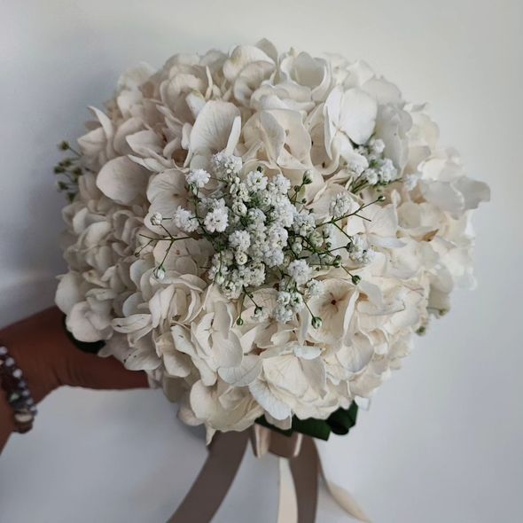 Bouquet da sposa bianco con ortensie e gypsophila, legato con un nastro su sfondo neutro.