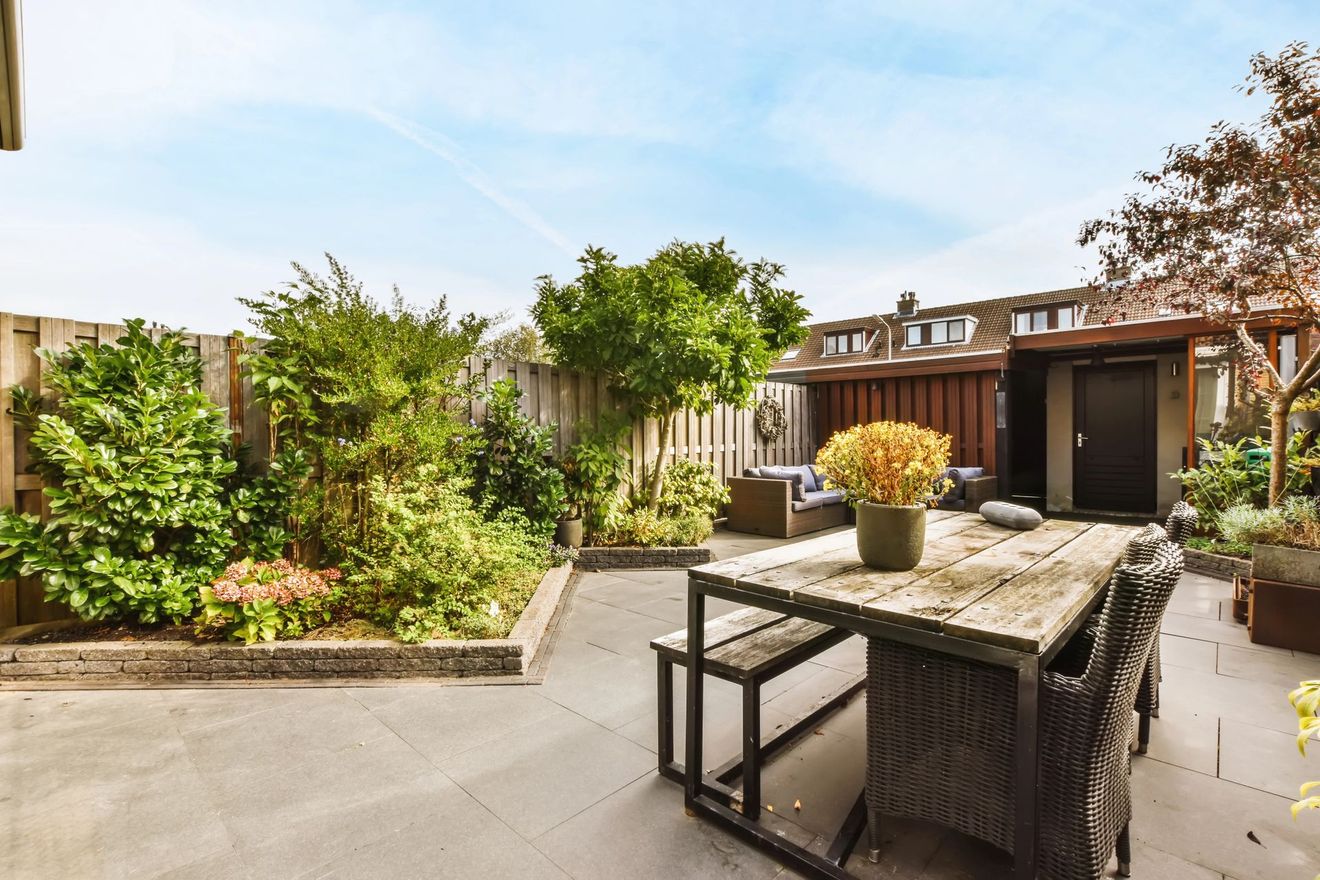 Patio soleggiato sul retro con aiuole rialzate, zona pavimentata con posti a sedere e tavolo da esterno con fiori in vaso.