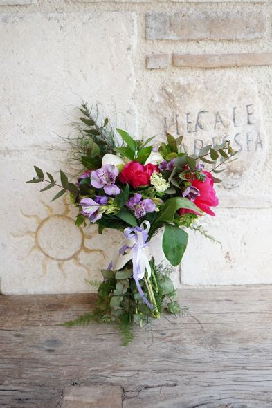 Bouquet di fiori rosa, viola, bianchi e rossi in un vaso trasparente, appoggiato a un muro di pietra.