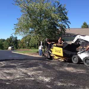 Asphalt paving in progress: workers operating machinery on a parking area with a tree and building in the background.