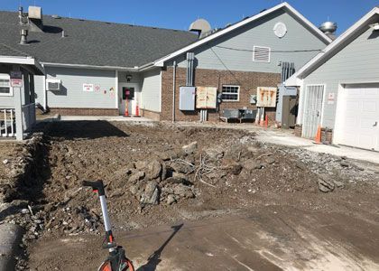 Construction site with excavated ground in front of a building. Brown dirt, grey buildings, clear sky.