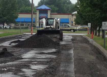 Bobcat spreading asphalt on a road in front of a building with blue awnings; cones and caution tape.