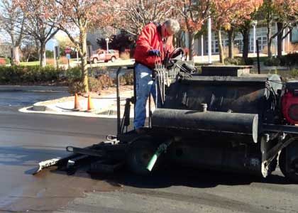 Person in red jacket operates asphalt sealing machine on a paved road.