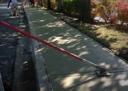 A worker using a long-handled tool to level wet concrete for a sidewalk.