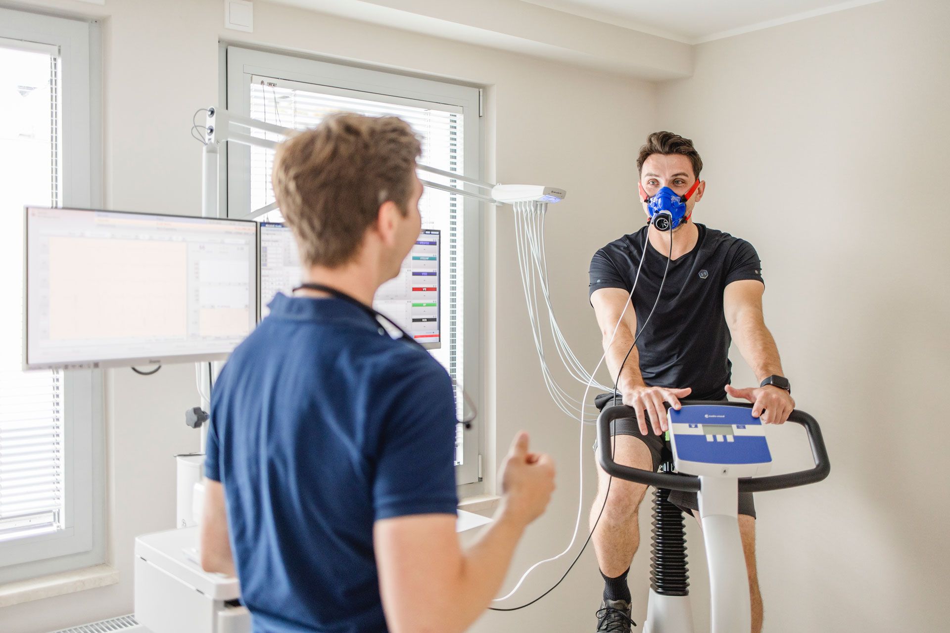 Mann mit einer Atemmaske auf einem Fahrrad, EKG-Fahrrad, verbunden mit Computer, Arzt schaut auf die Ergebnise am Computer, Spiroergometrie