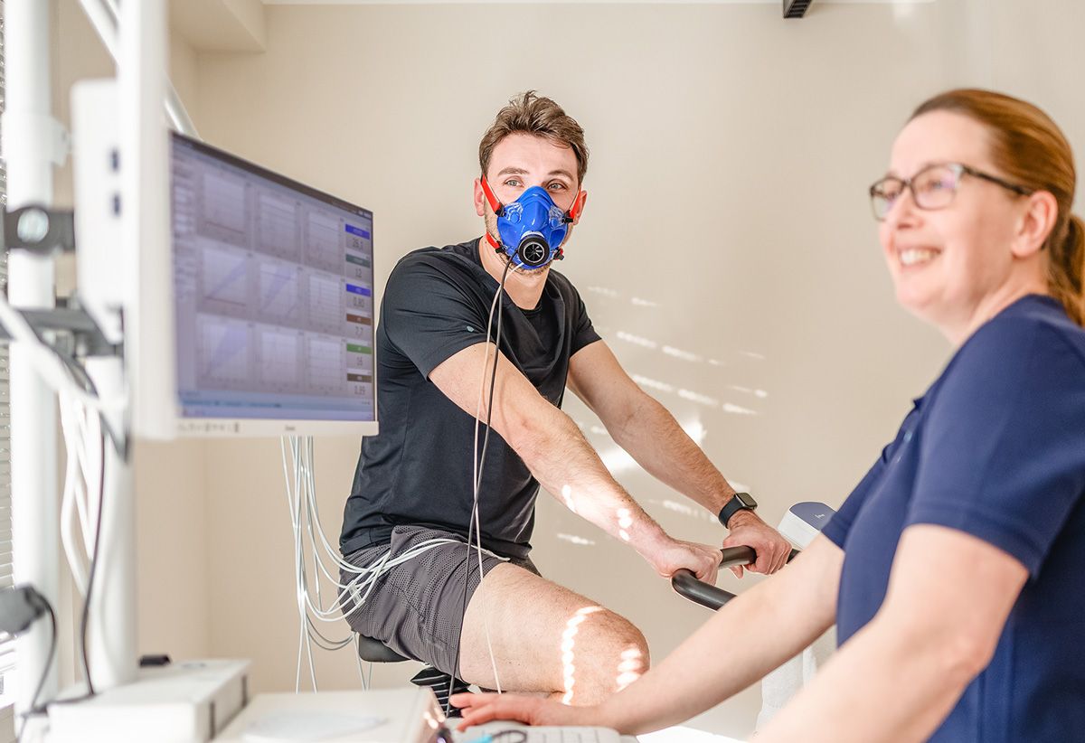 Mann mit einer Atemmaske auf einem Fahrrad, EKG-Fahrrad, Arzt schaut auf die Ergebnise am Computer, Spiroergometrie