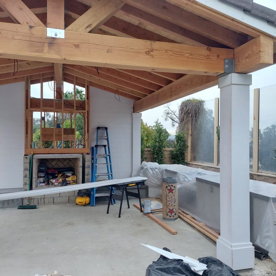 Construction of an outdoor patio with wooden beams, fireplace, and white pillars; tools and materials scattered around.