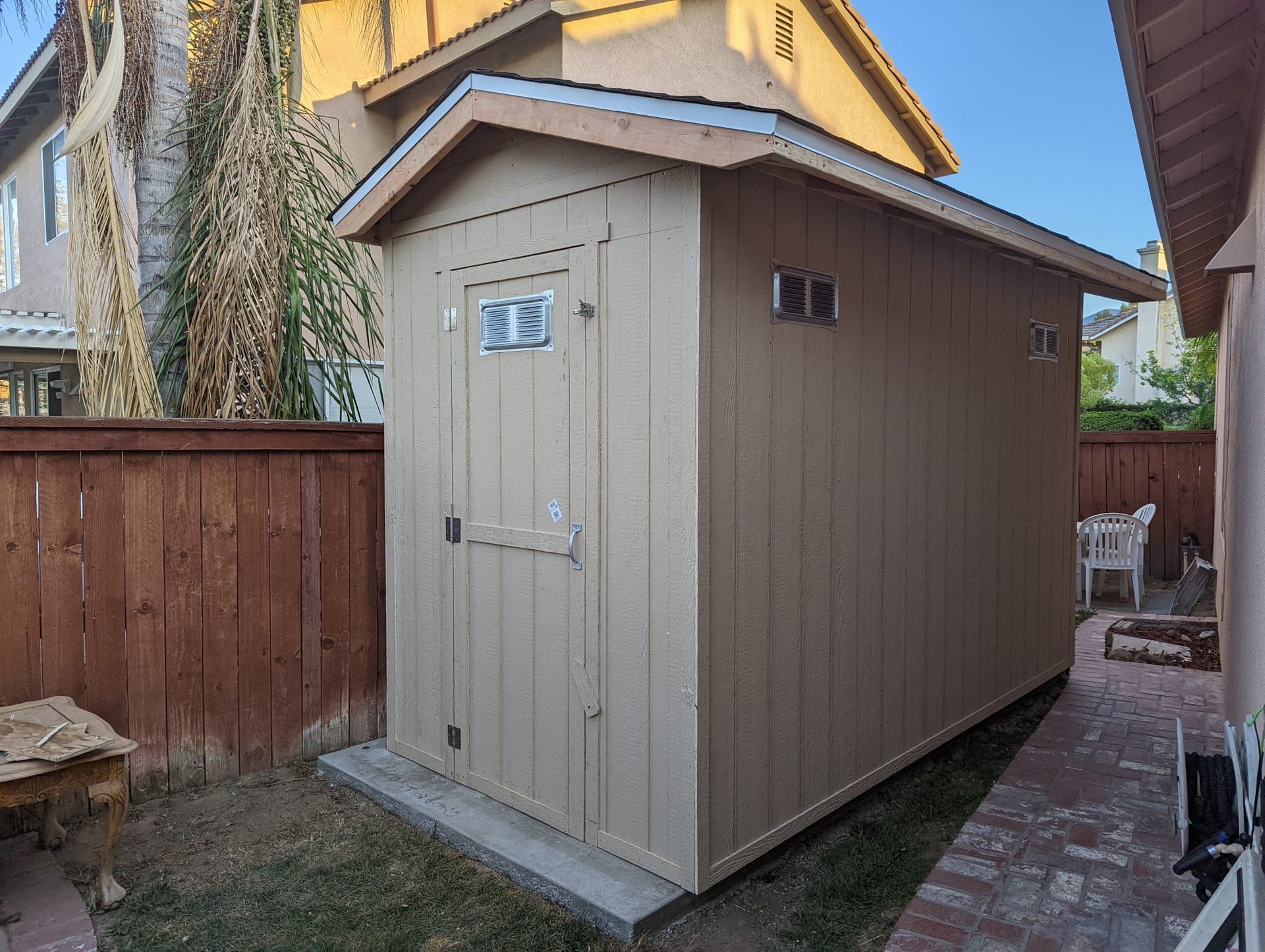 Tan storage shed in a backyard with a wooden fence.
