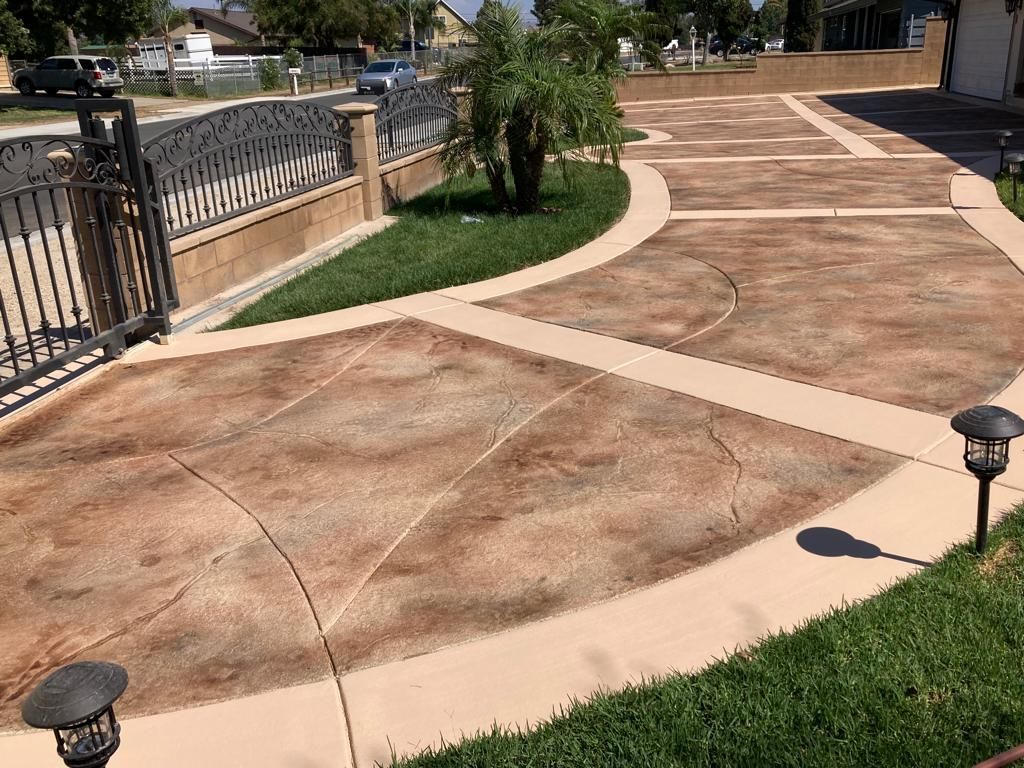 Decorative concrete driveway with tan borders and brown textured panels, edged by green grass and a black gate.