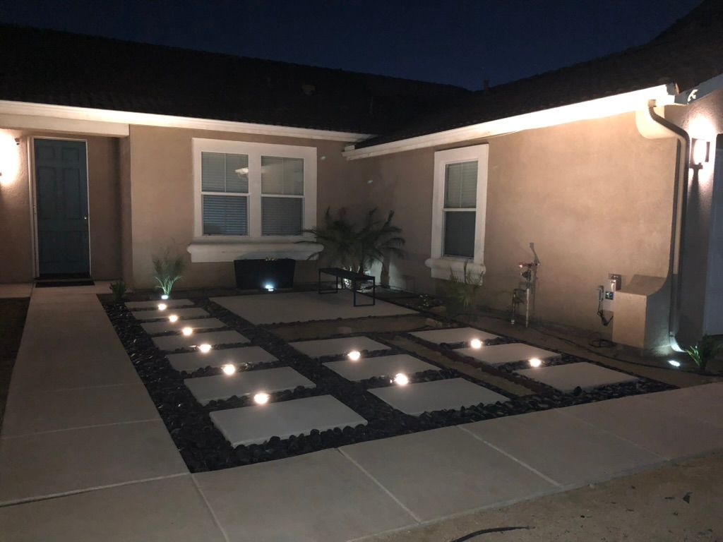 Night view of a modern patio with lit stone pavers, black gravel, and built-in lighting on a tan-colored house.