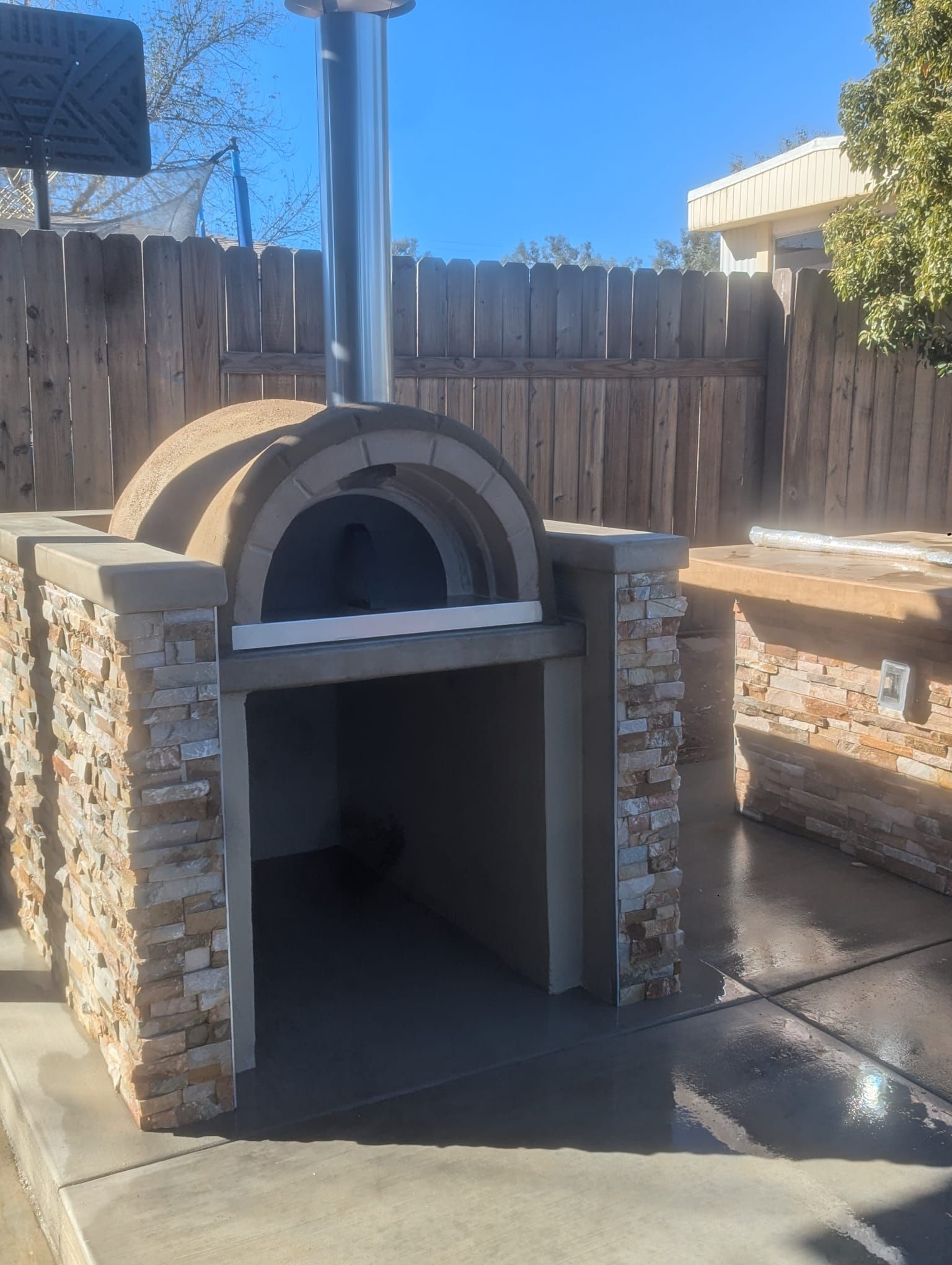 Outdoor pizza oven with stone facade, chimney, and adjacent countertop, against a wooden fence.