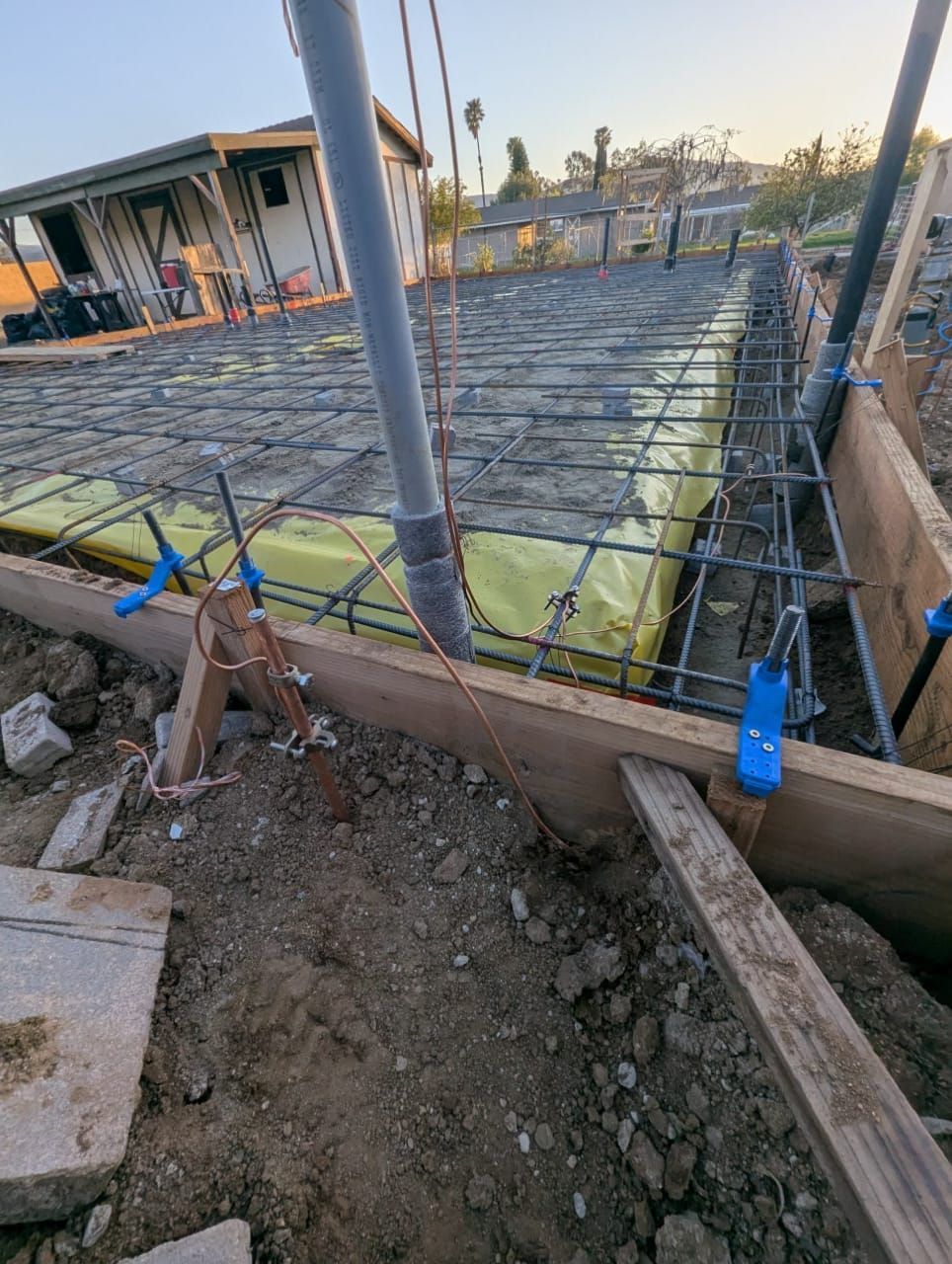 Concrete foundation construction with rebar framework and plumbing pipes. Yellow insulation visible.