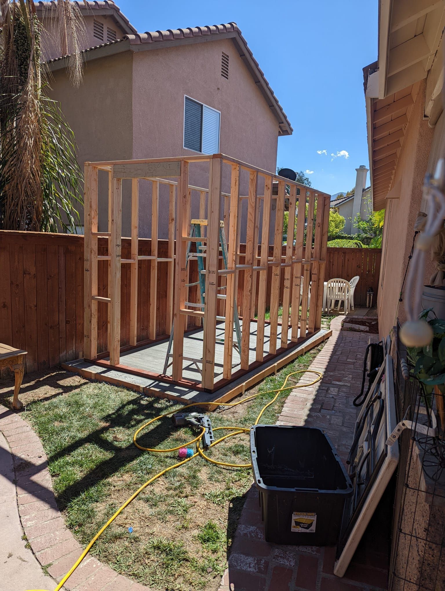 Wooden frame of a shed under construction in a backyard, with tools and a fence visible.