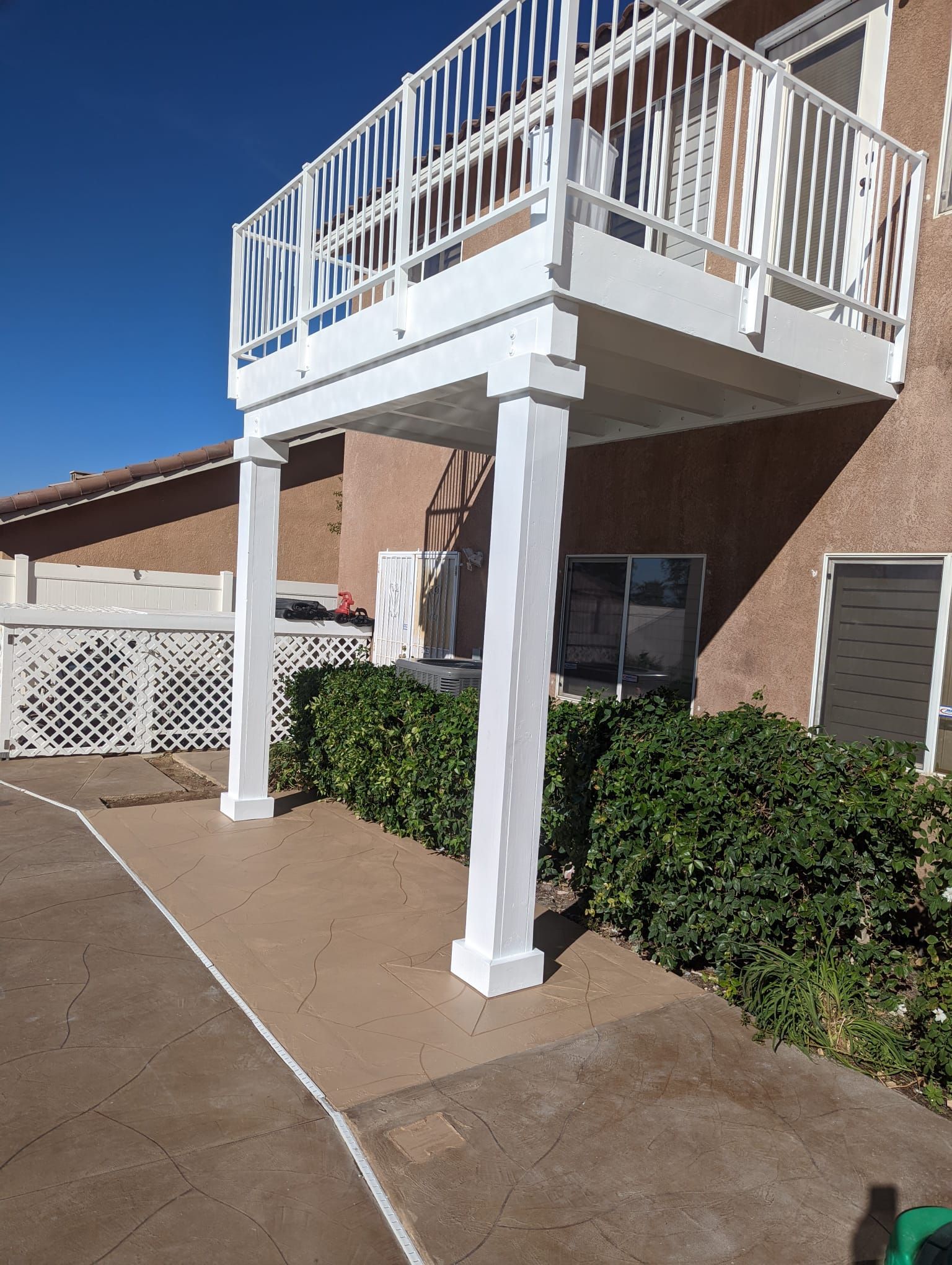 White balcony supported by columns, over concrete path and bushes, against building.