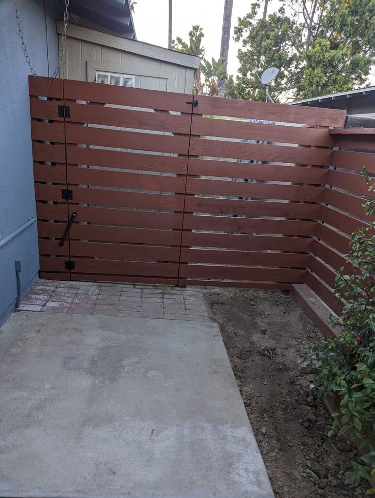 Brown wooden gate in a small, concrete-paved outdoor space, next to a blue wall and a leafy hedge.