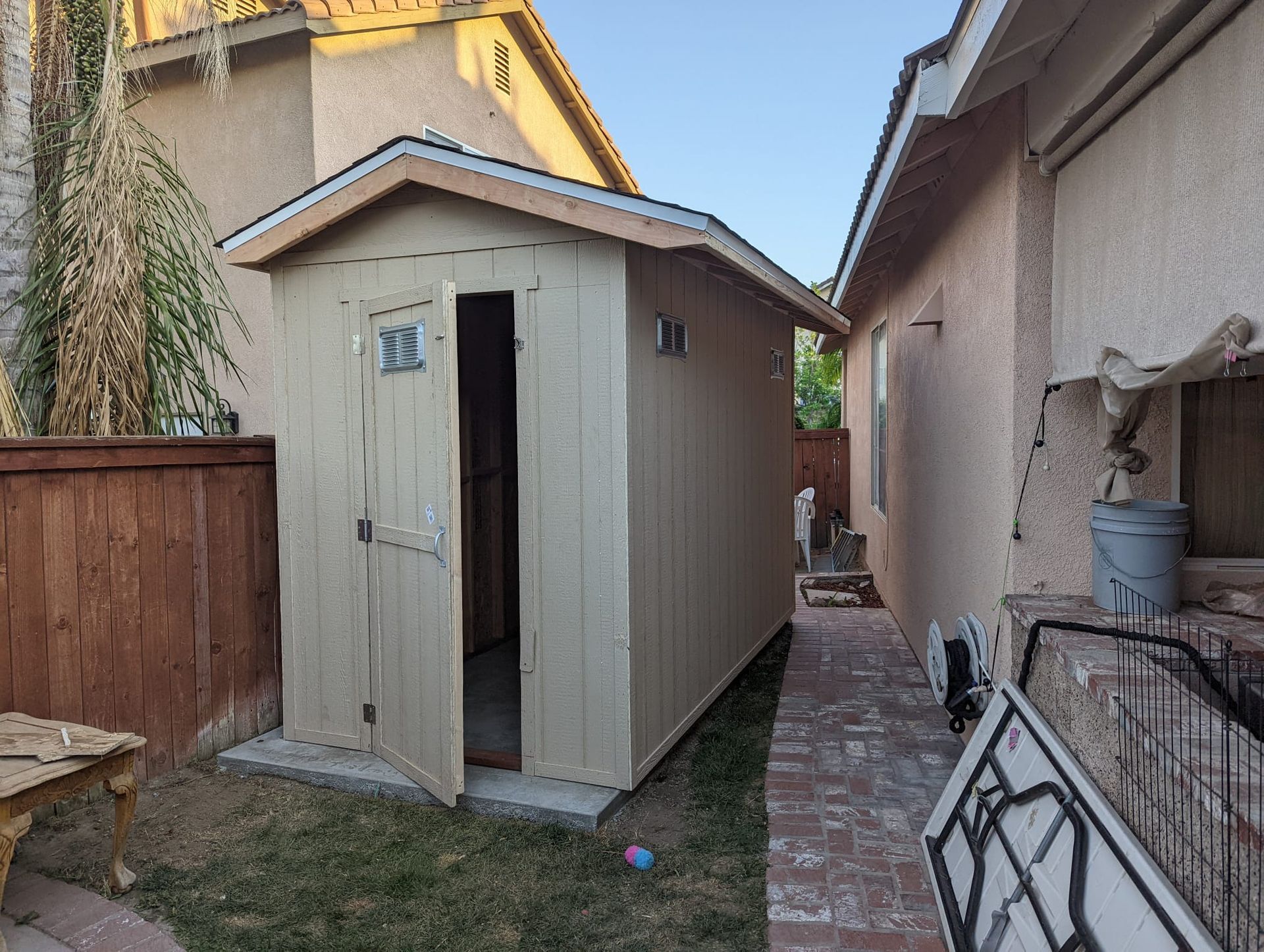 Tan shed with open door in a backyard, next to a house. Wooden fence and brick pathway visible.