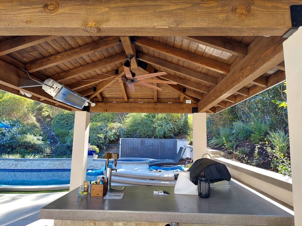 Patio with wooden beams, ceiling fan, and outdoor bar overlooking a pool and foliage.