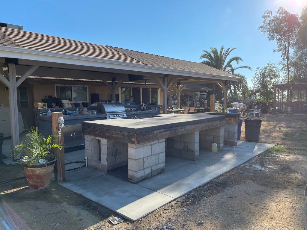 Outdoor kitchen with concrete block base, wooden countertop, grill, under a covered patio.