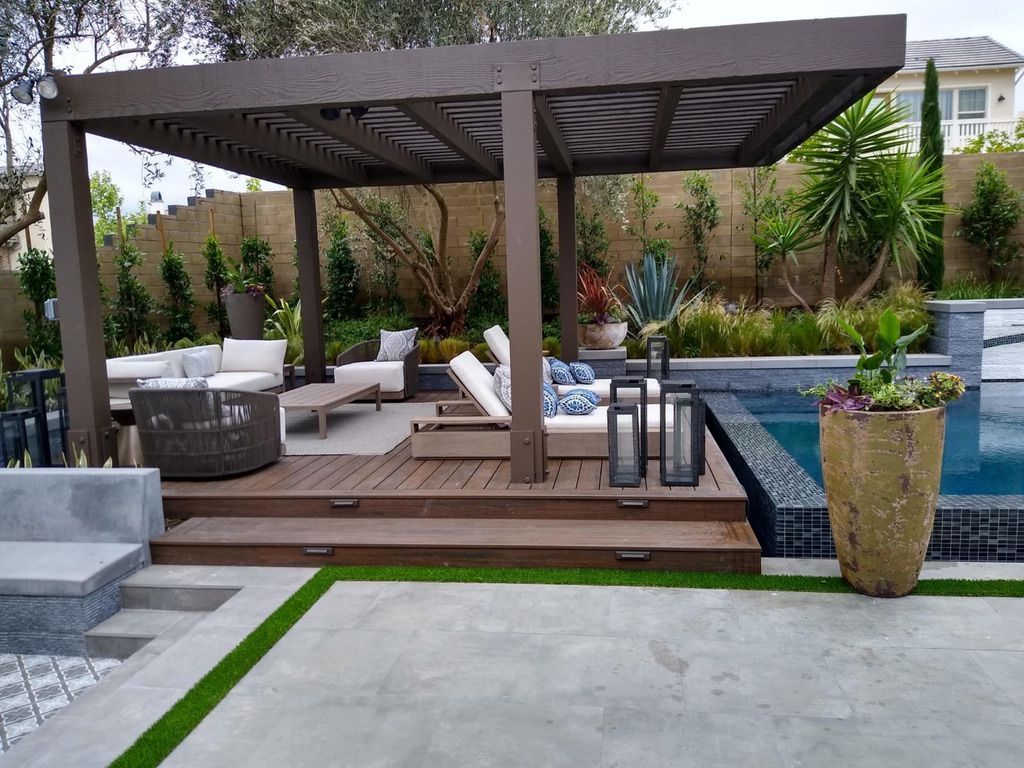 Outdoor lounge area with a brown pergola, pool, deck, and seating. Lush greenery and modern design.