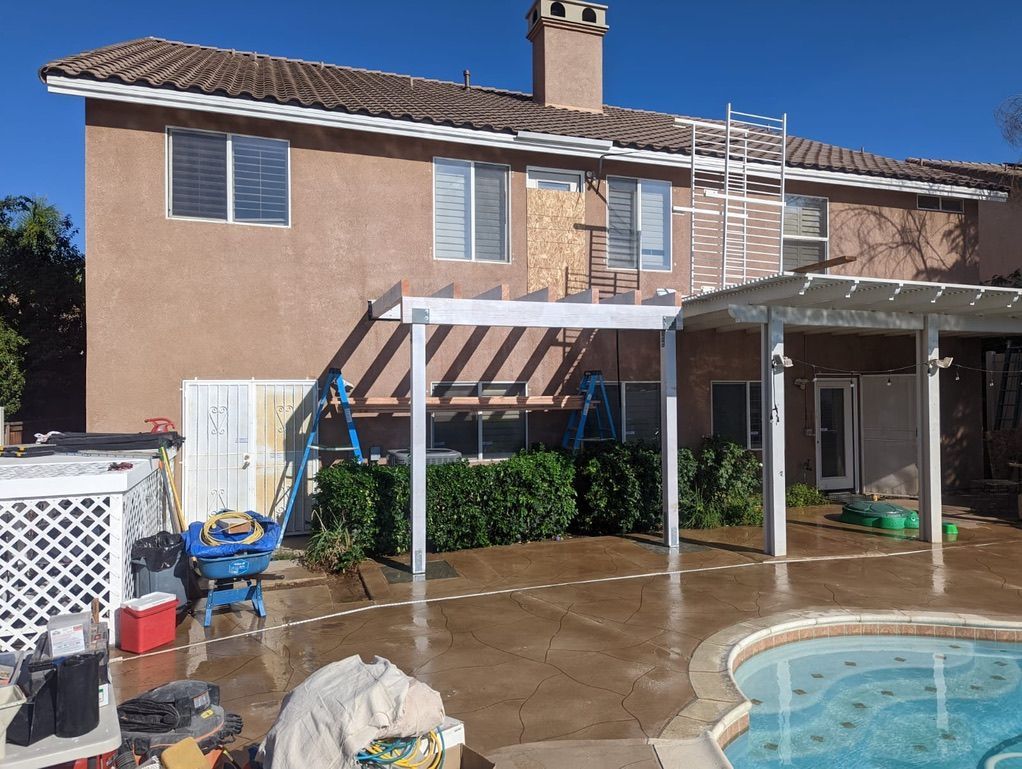 Two-story house with a backyard pool, pergola, and exterior work in progress; tan walls, brown roof, blue sky.