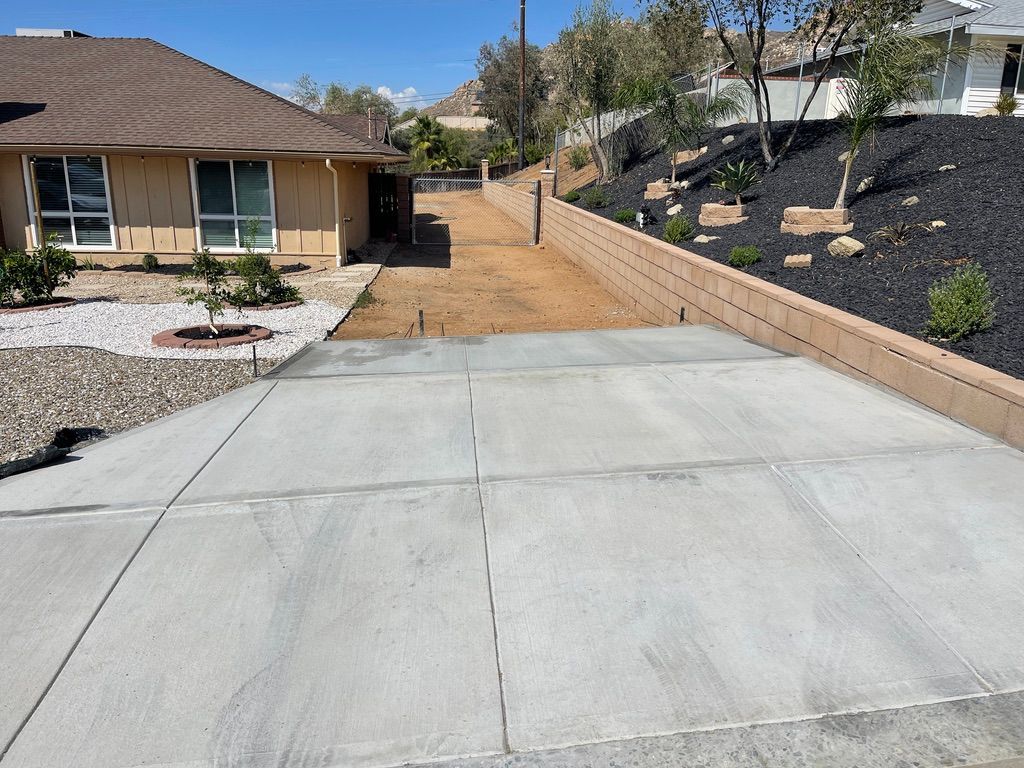 Concrete driveway leading to a house with a brown roof and a dirt path. Black mulch and a retaining wall on the right.