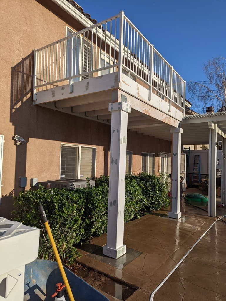 White deck elevated above bushes, supported by white columns. Tan house, blue sky.