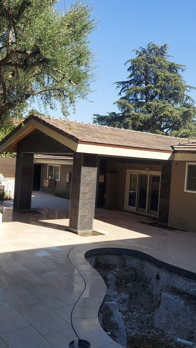 Patio with pool, covered walkway with stone columns, beige house, and trees under a clear blue sky.