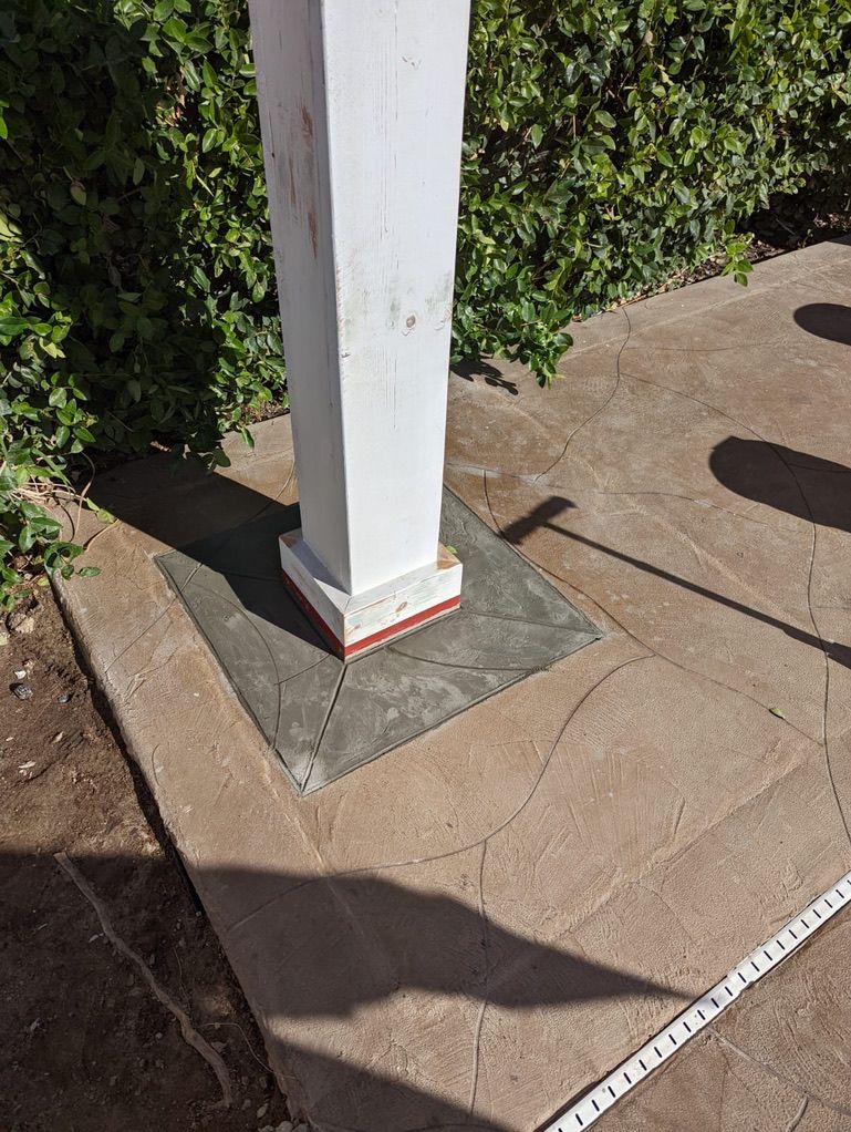 White porch column on a concrete base, set on a textured patio; greenery in the background.