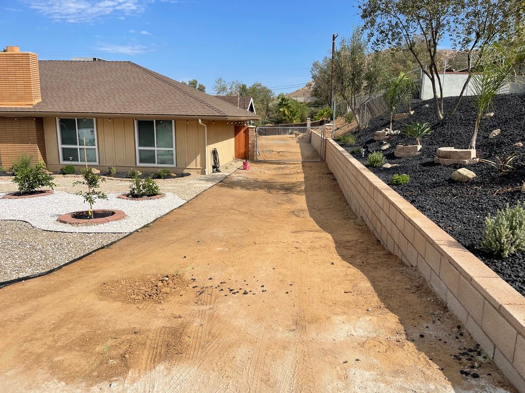 A dirt driveway next to a retaining wall and house with tan stucco and a brown roof.