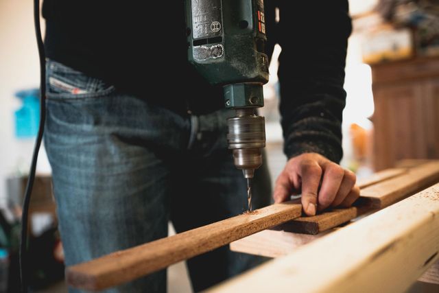 Person drilling a hole in a wooden board with an electric drill, wearing jeans and a black sweatshirt.
